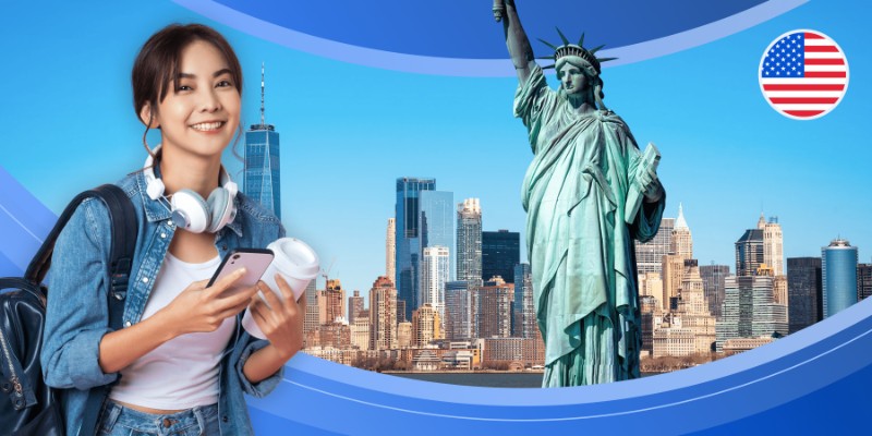 A smiling young woman with headphones around her neck, holding a smartphone and a coffee cup, set against a backdrop of the New York City skyline and the Statue of Liberty. The image features a blue curved border and a circular U.S. flag icon in the top right corner, symbolizing the experience of an international student studying in the United States.