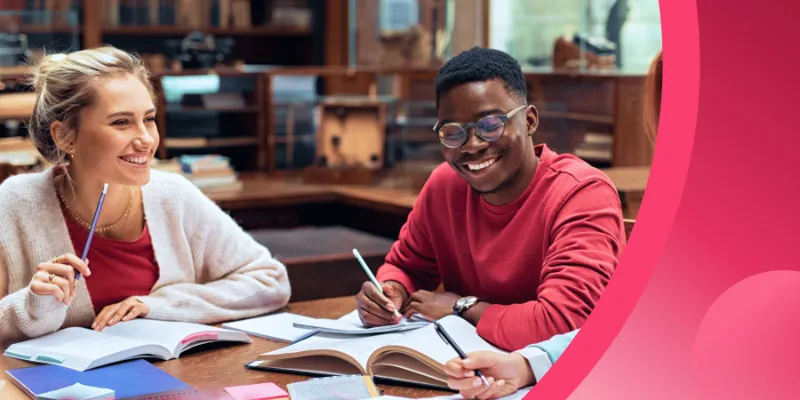 Two smiling students, a woman and a man, studying together with open books at a table in a library