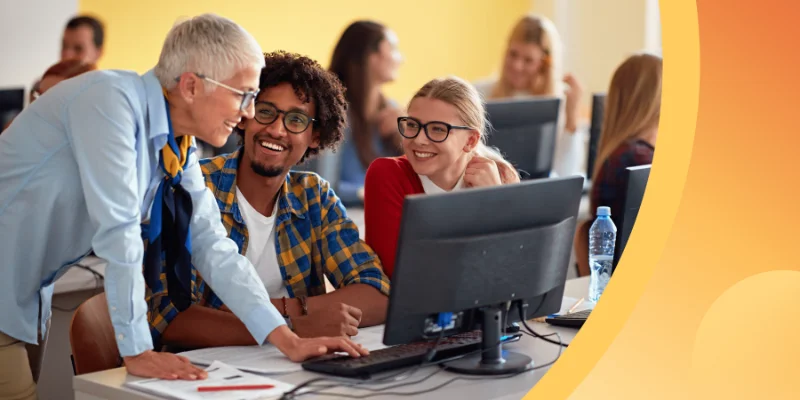 An instructor with short grey hair leans over a desk to assist two smiling adult students working at a computer terminal in a bright classroom.
