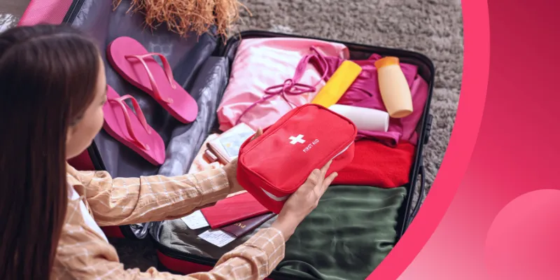 A young woman packing an open pink suitcase on the floor, placing a red first-aid kit on top of neatly folded clothes, toiletries, flip-flops, and travel documents.