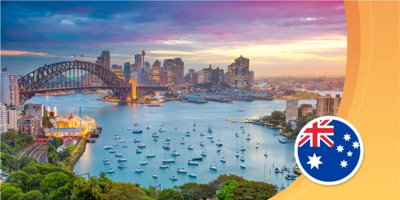 Panoramic view of the Sydney Harbour Bridge and city skyline at sunset, with boats moored in the harbour and a circular Australian flag icon in the bottom right corner.